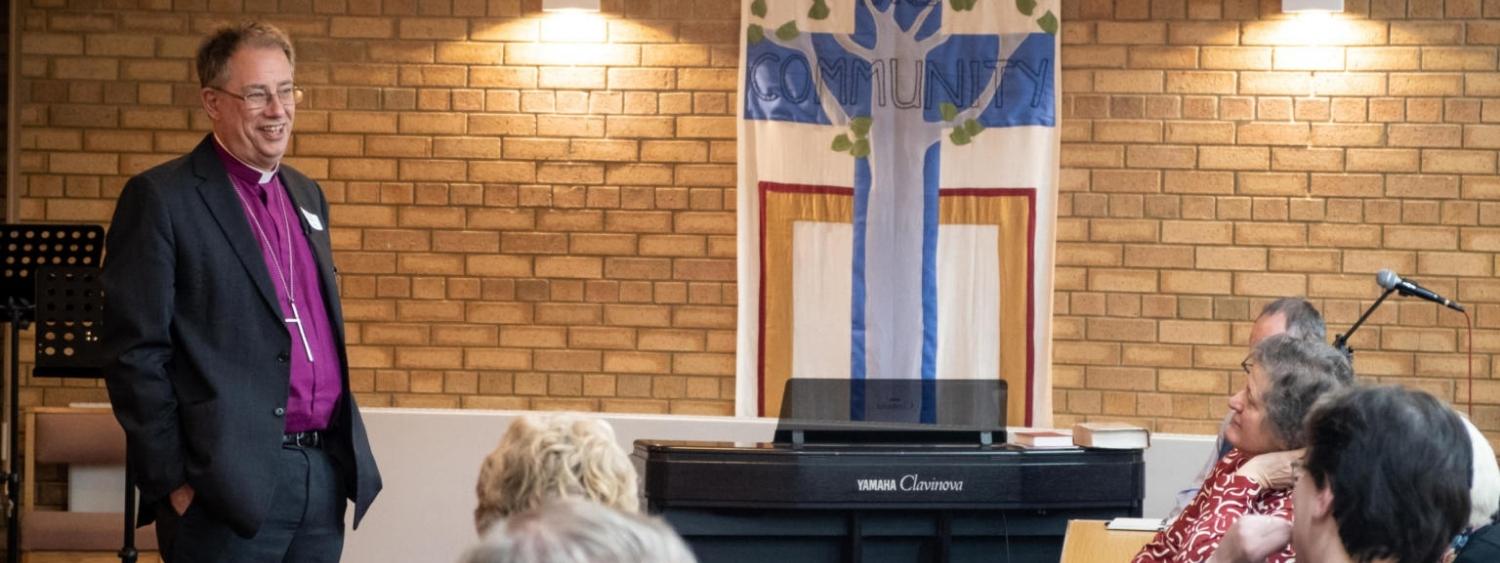 The Bishop of Oxford stands smiling talking to PCC members seated in a church hall with a brick wall background