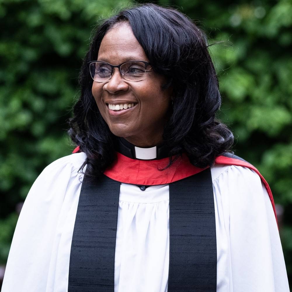 The Revd Canon Janet Binns smiling and looking to the left in front of green shrubs